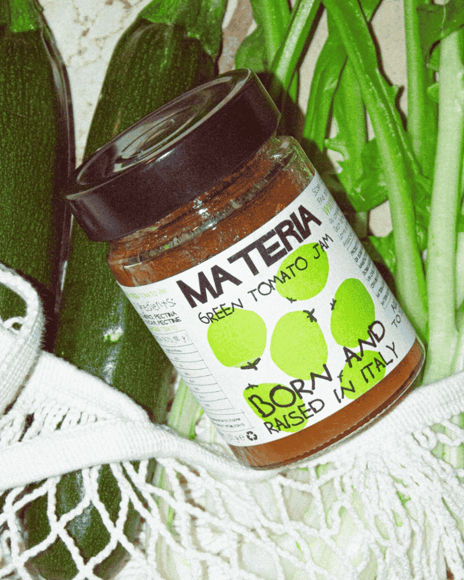 Glas Materia Green Tomato Jam auf frischem Gemüse, darunter Zucchini und Frühlingszwiebeln, in einem Netzbeutel.