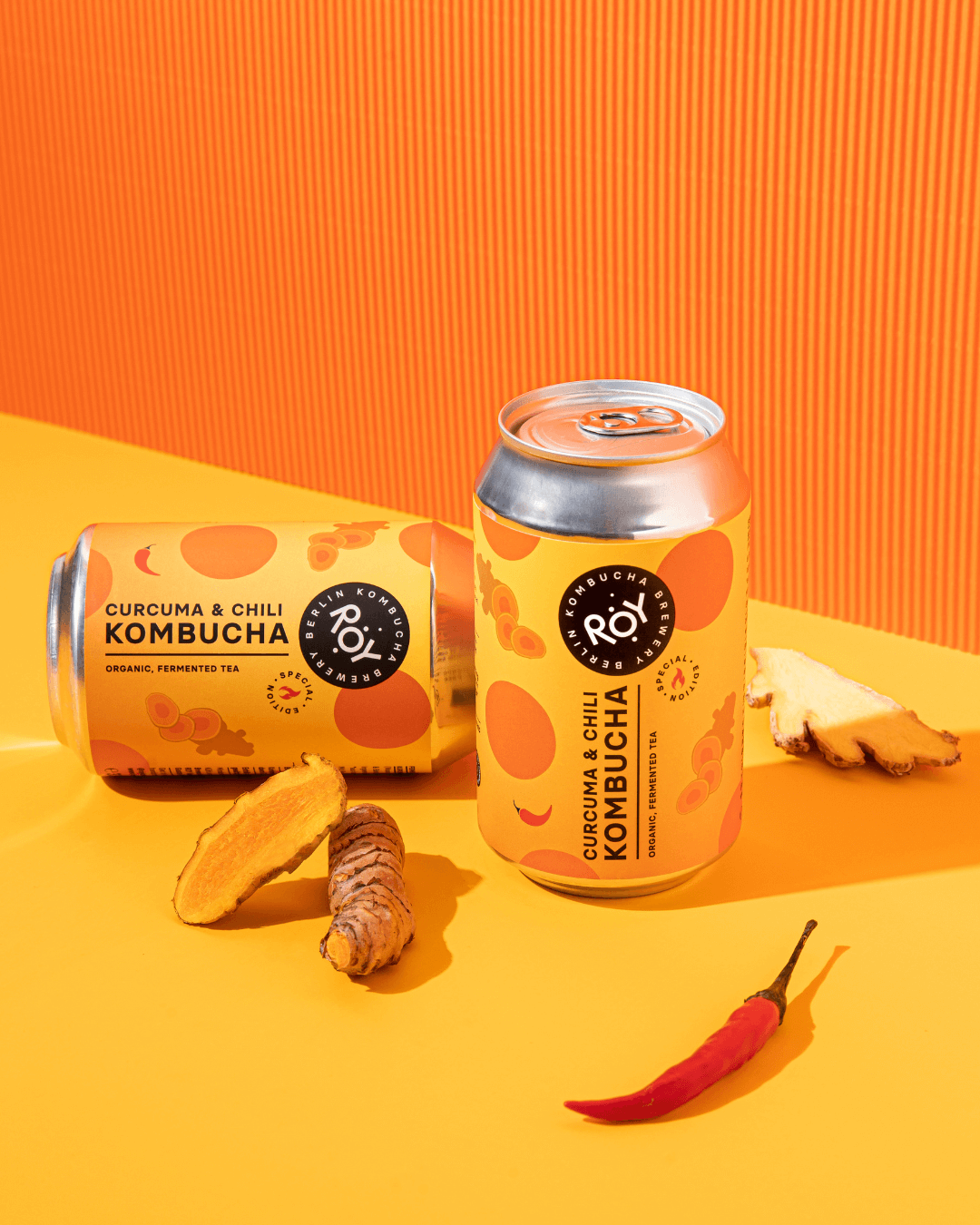 Roy Bio-Kombucha Curcuma & Chili, inszeniert mit frischer Kurkuma und roter Chili auf leuchtendem Orange.
