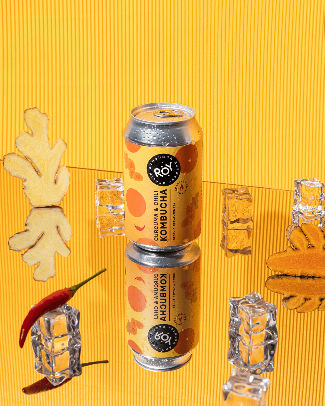 ROY Bio-Kurkuma-Chili-Kombucha stilvoll inszeniert mit Ingwer, Chili, Eiswürfeln und Spiegelung auf gelbem Hintergrund.