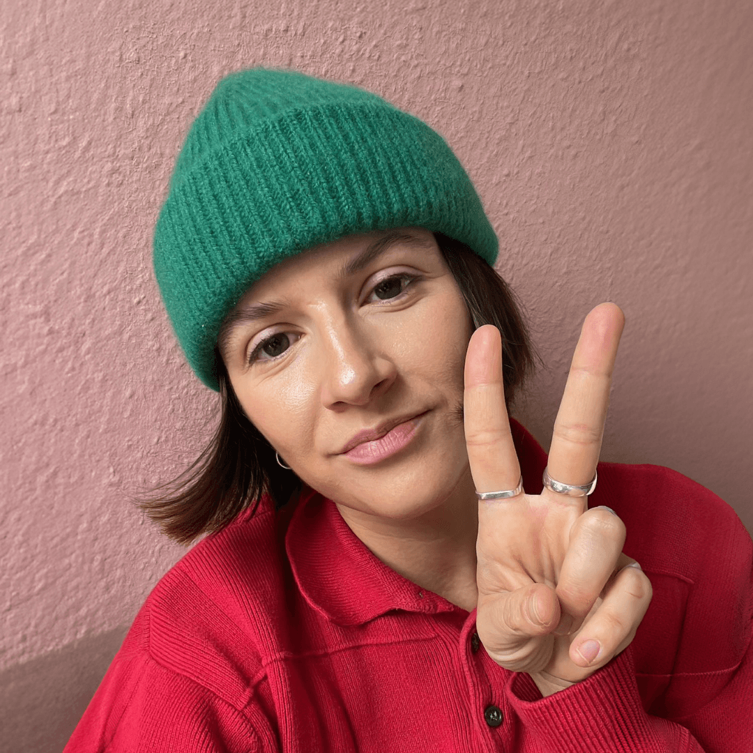 Person mit grüner Strickmütze und rotem Pullover zeigt ein Peace-Zeichen vor einer rosa Wand.