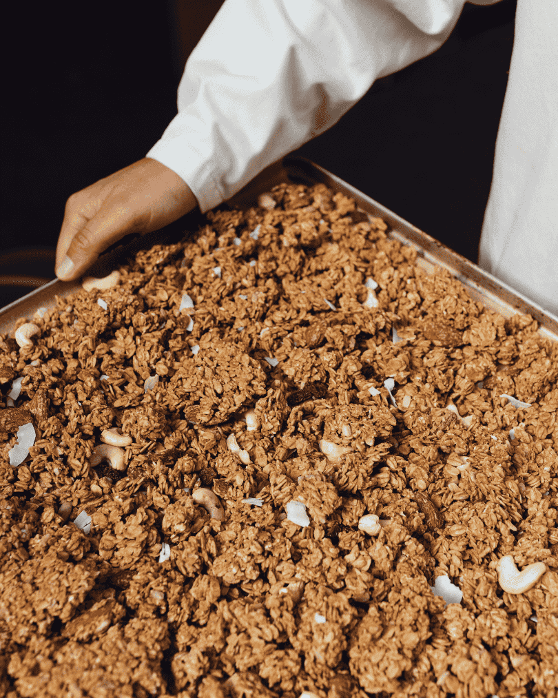 Frisches knuspriges Granola mit Nüssen und Kokos auf einem Blech, gehalten von einer Person in weißer Kleidung.