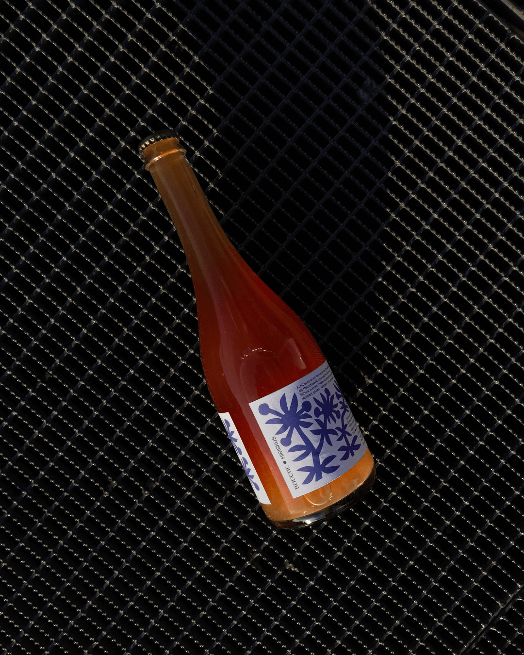 Eine Flasche Hibiskus-Pet-Nat-Kombucha auf einem schwarzen Gitterhintergrund, mit einem blauen Etikett.