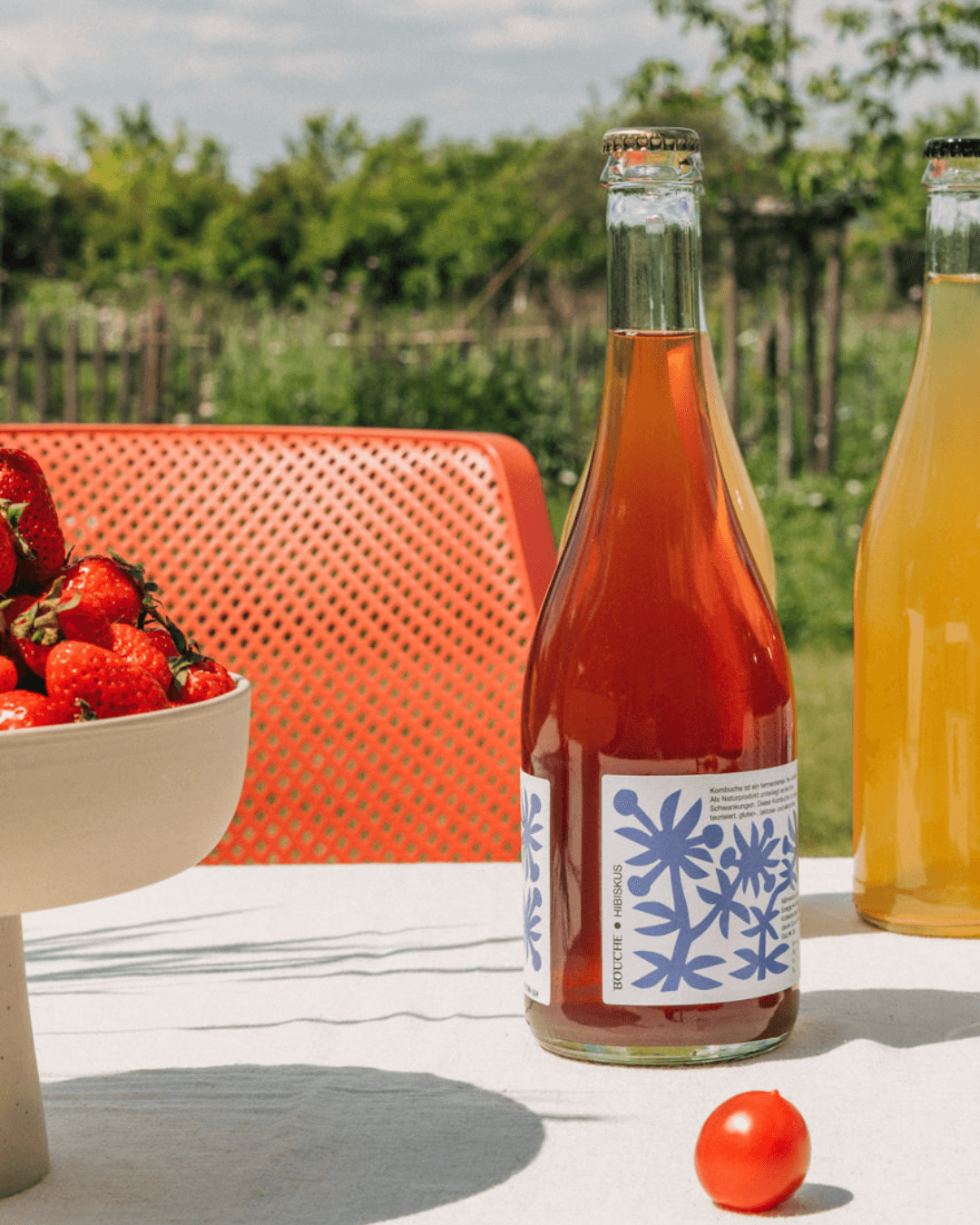 Zwei Flaschen Hibiskus-Pet-Nat-Kombucha auf einem Tisch mit roten Erdbeeren und einer Tomate, im Hintergrund grünes Gras.