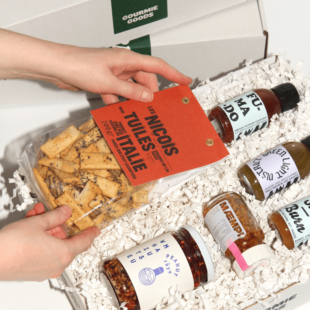Hand legt feine Cracker in eine Gourmet-Geschenkbox von Gourmie Goods mit internationalen Delikatessen und Saucen.
