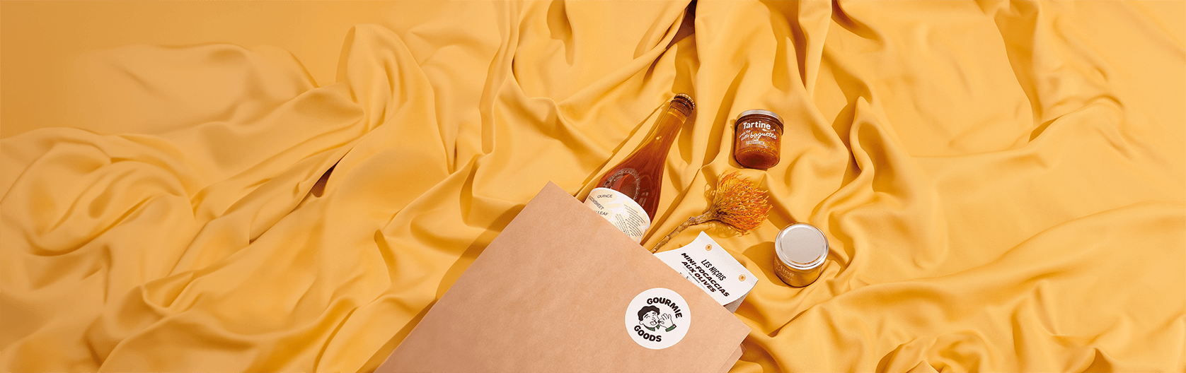 Geschenkbox von Gourmie Goods mit hochwertigen Produkten, darunter eine Flasche, ein Glas Marmelade, ein Blumenakzent und weitere Spezialitäten, präsentiert auf einem goldgelben Stoffhintergrund.