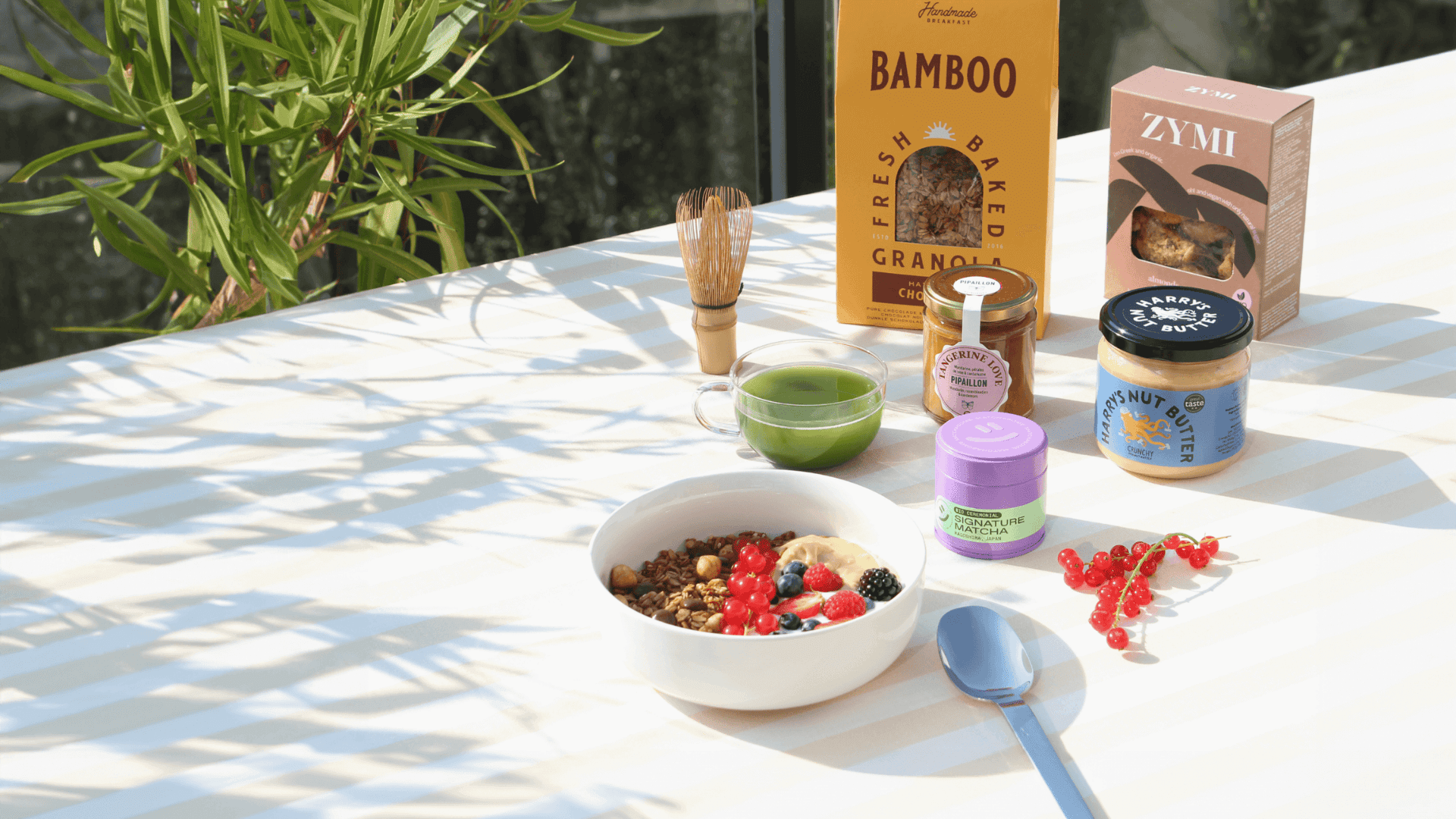 Ein sonniger Frühstückstisch mit Granola-Bowl, Beeren, Matcha-Tee, Nussbutter, Honig und Gebäck, umgeben von Licht.