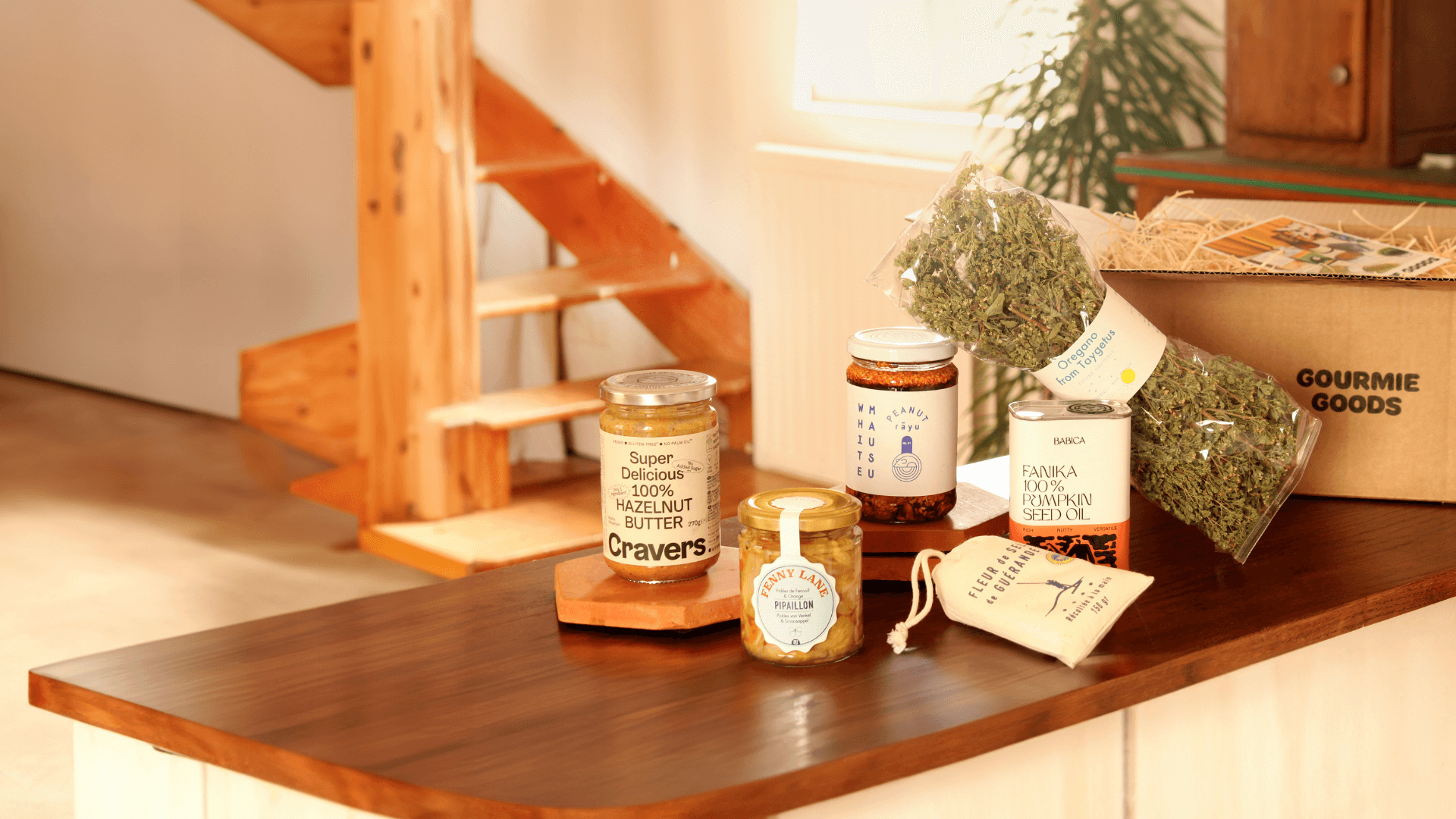 Feinkostprodukte wie Haselnussbutter, Kürbiskernöl, Oregano auf einer Holztheke, mit einer Gourmie Goods Box im Hintergrund.