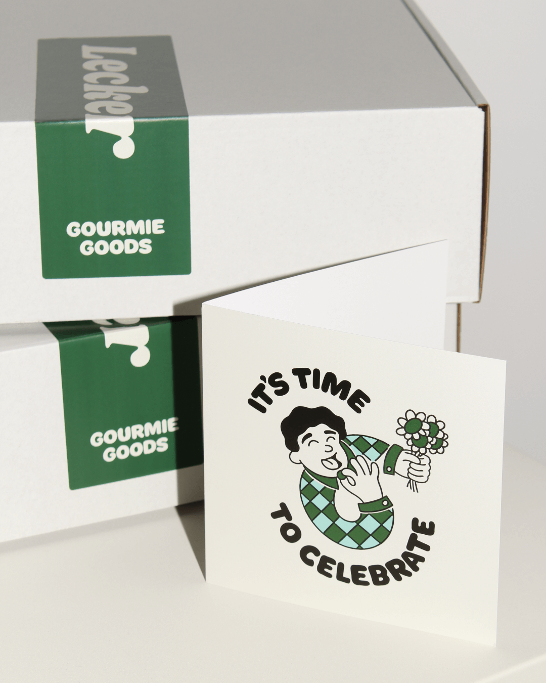 Eine Grußkarte mit der Aufschrift „It's time to celebrate“ liegt vor zwei Gourmie Goods Geschenkboxen auf einem Tisch.