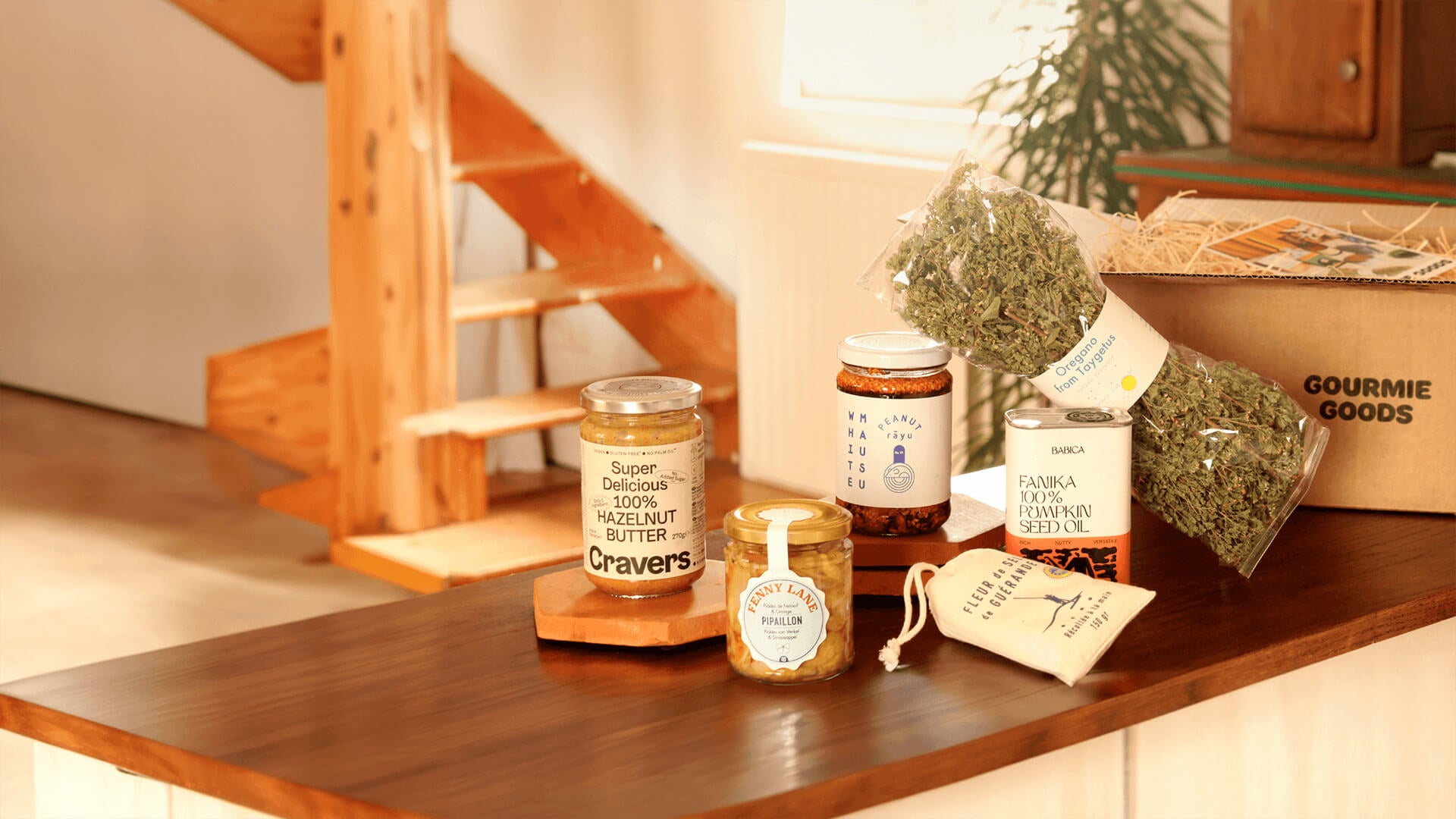 Feinkostprodukte wie Haselnussbutter, Kürbiskernöl, Oregano auf einer Holztheke, mit einer Gourmie Goods Box im Hintergrund.