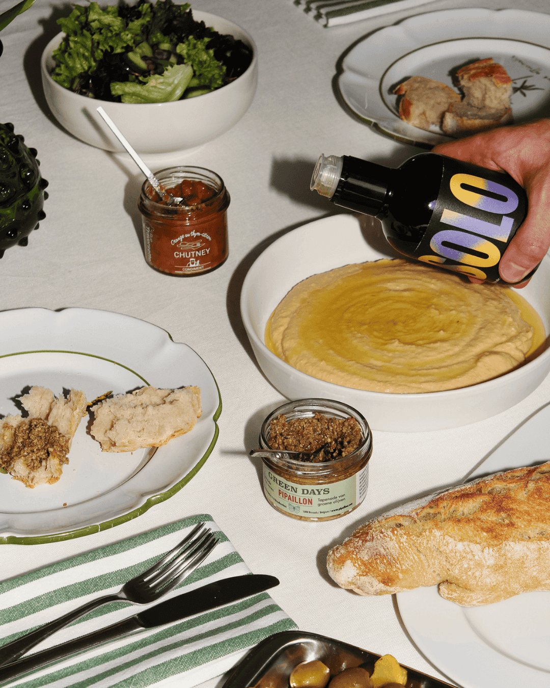 Ein Tisch mit mediterranen Speisen, darunter Hummus, Olivenöl, Brot, Salat, Chutney und Tapenade, auf weißer Tischdecke.