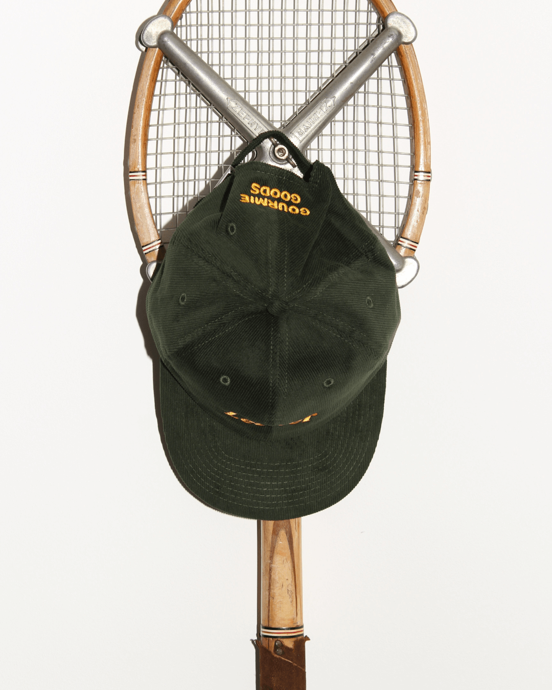 Dunkelgrüne Kord-Kappe von Gourmie Goods mit gelbem „Gourmie Goods“-Stick, aufgehängt an einem Vintage-Tennisschläger.