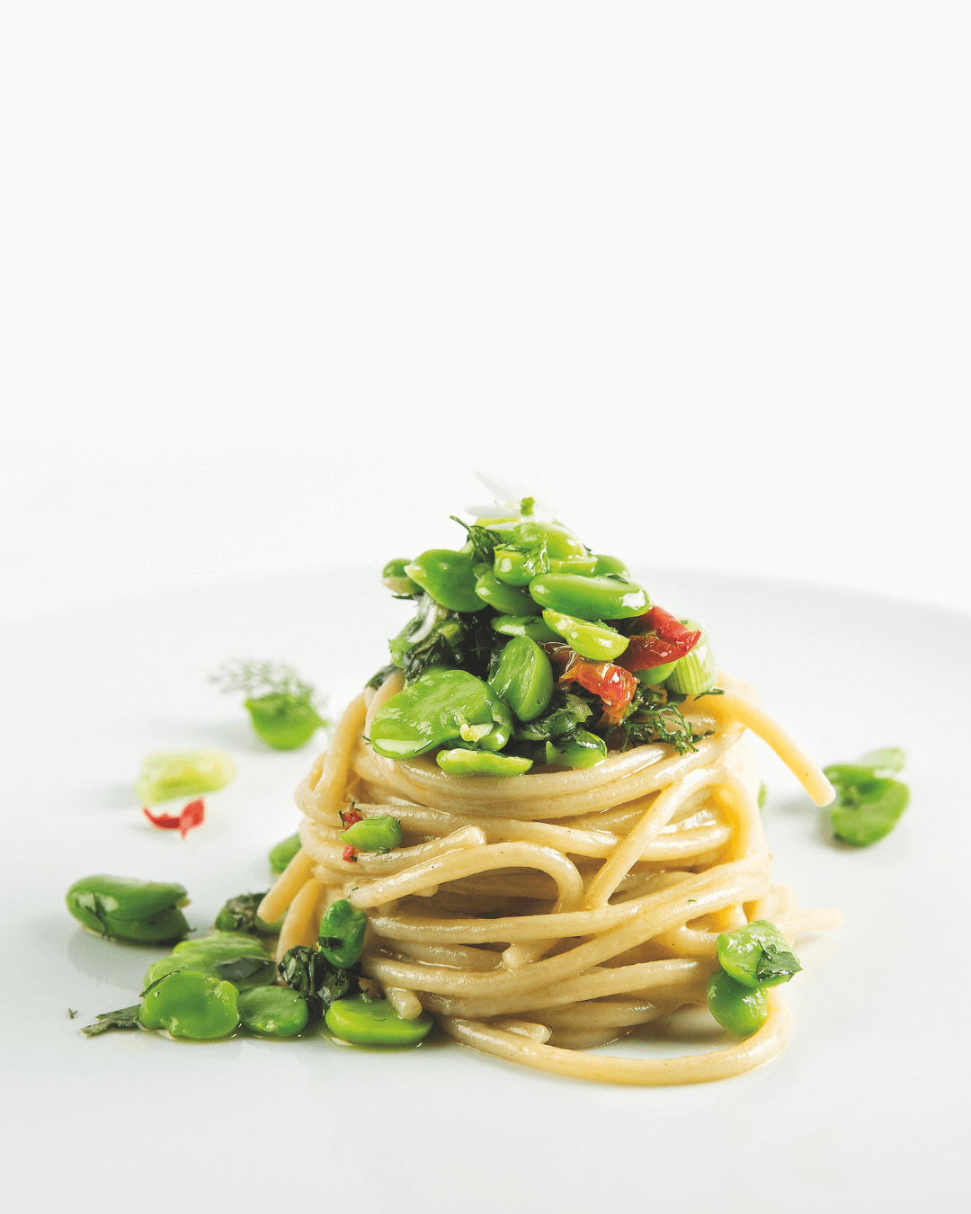 Spaghetti-Nest mit dicken Bohnen, frischen Kräutern und Chili auf weißem Teller.