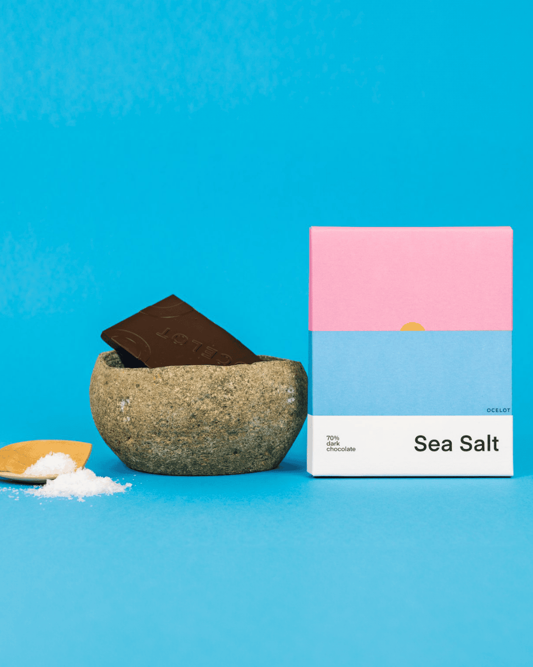 Schokoladentafel "Sea Salt" von Ocelot neben einer Schale mit Salz und einer Stück Schokolade auf blauem Hintergrund.