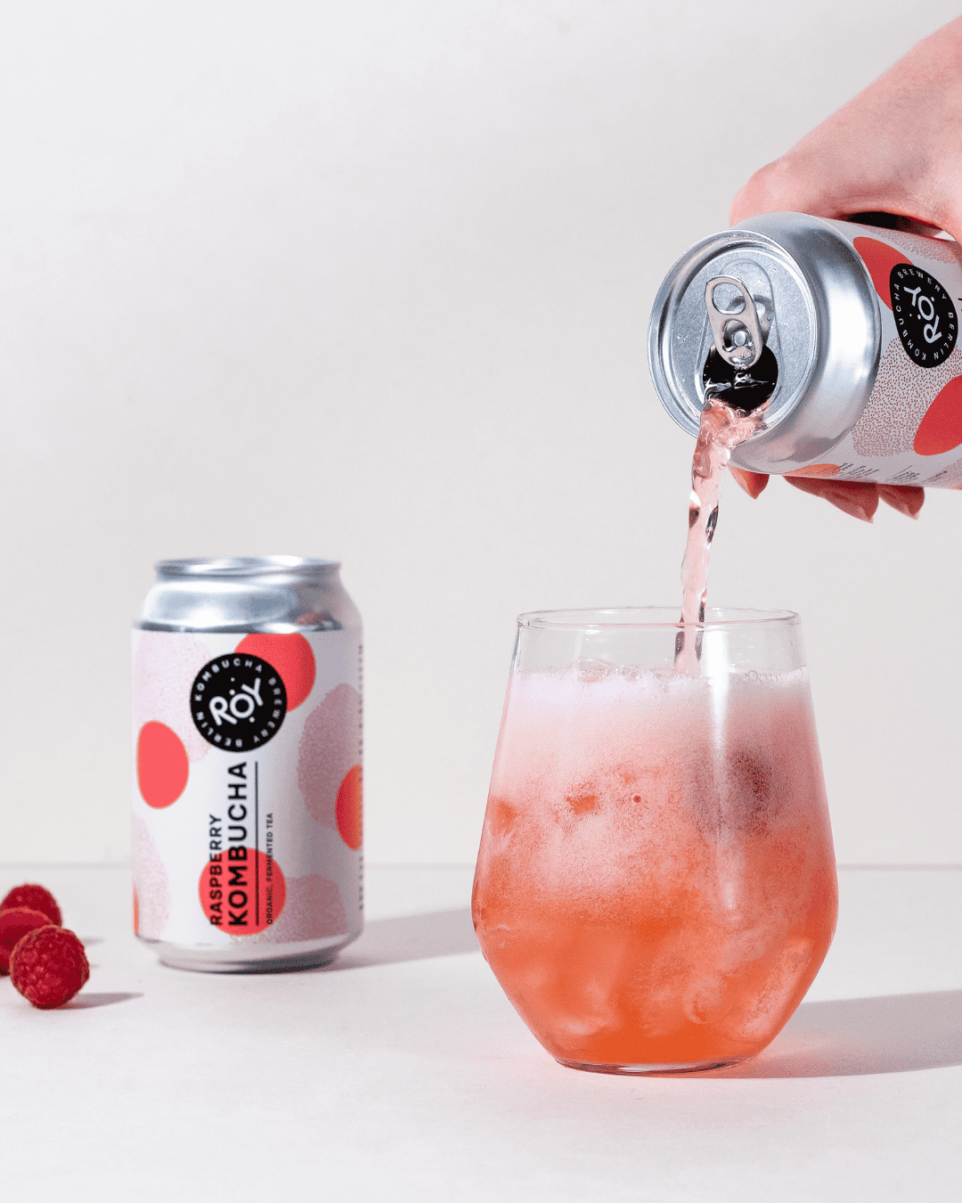 Hand gießt ROY Bio-Himbeer-Kombucha in ein Glas mit Eiswürfeln ein, daneben eine weitere Dose und frische Himbeeren.