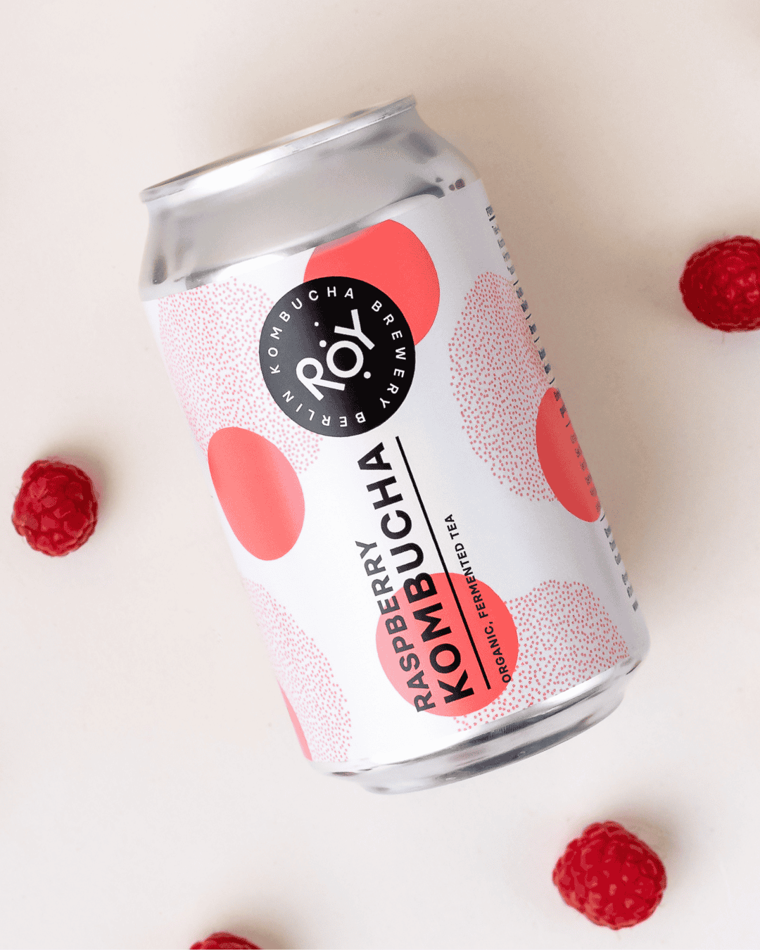 ROY Bio-Himbeer-Kombucha-Dose mit Polka-Dot-Design, auf hellem Hintergrund mit verstreuten frischen Himbeeren arrangiert.