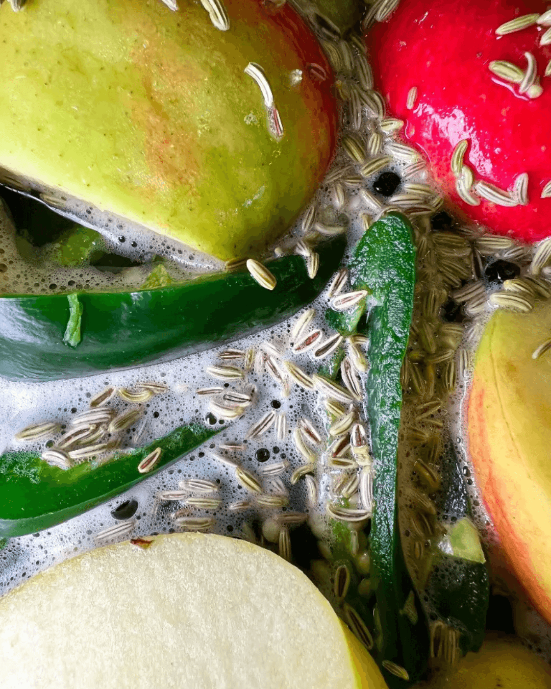 Geschnittene Äpfel, grüne Chilis und Gewürze wie Fenchelsamen in Flüssigkeit, vorbereitet zur Fermentation.